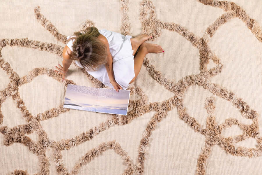 Woven For Him rug by Moe Khadra, featuring subtle Arabic calligraphy and a natural palette, with a model sitting on it.