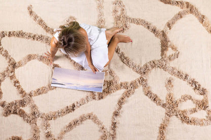 Woven For Him rug by Moe Khadra, featuring subtle Arabic calligraphy and a natural palette, with a model sitting on it.