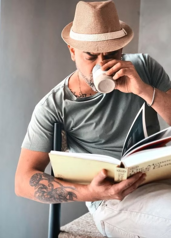 Man drinking from a cup and reading a book with a neutral background